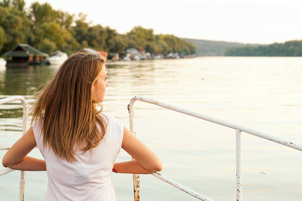 Beautiful young woman is looking at sunset over the river Attractive woman enjoys summer and warm sunlight. She is admiring beautiful view and enjoying sunset sunlight. The wind blew her hair.