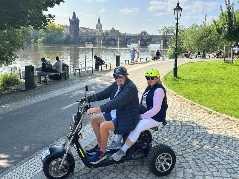 Paar auf einem Adventure Trike Elektroroller mit der Karlsbrücke und der Moldau im Hintergrund, Prag